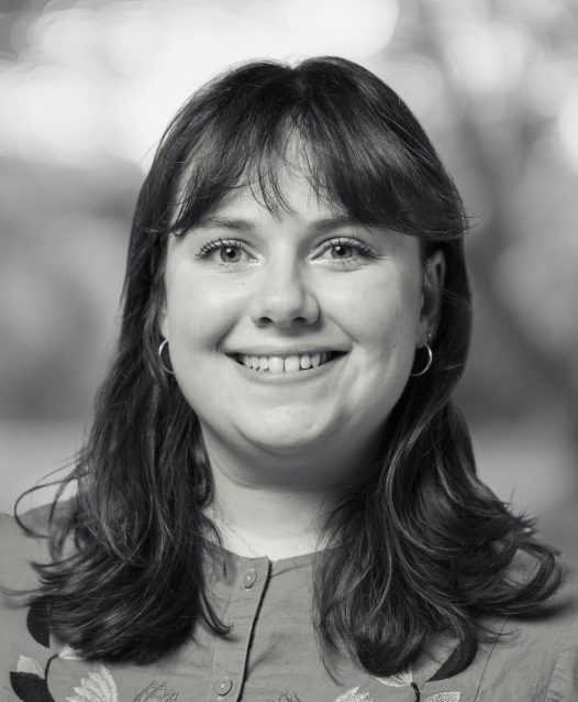 A person with shoulder-length hair smiles warmly in a black-and-white portrait, with a blurred background of trees and light.