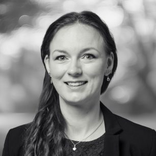A woman with long hair, wearing a blazer and earrings, smiles in a black and white portrait with a blurred background.
